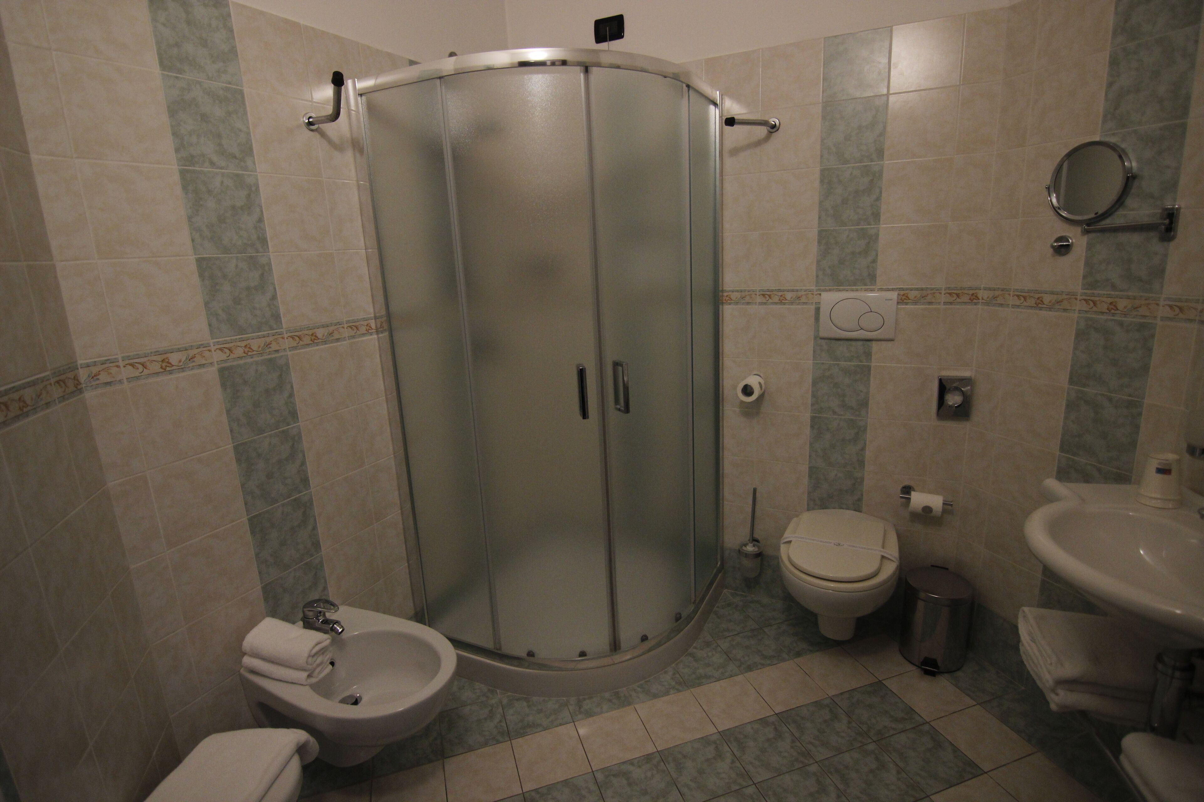 A bathroom with a shower enclosure, toilet, bidet, sink, and a trash can.