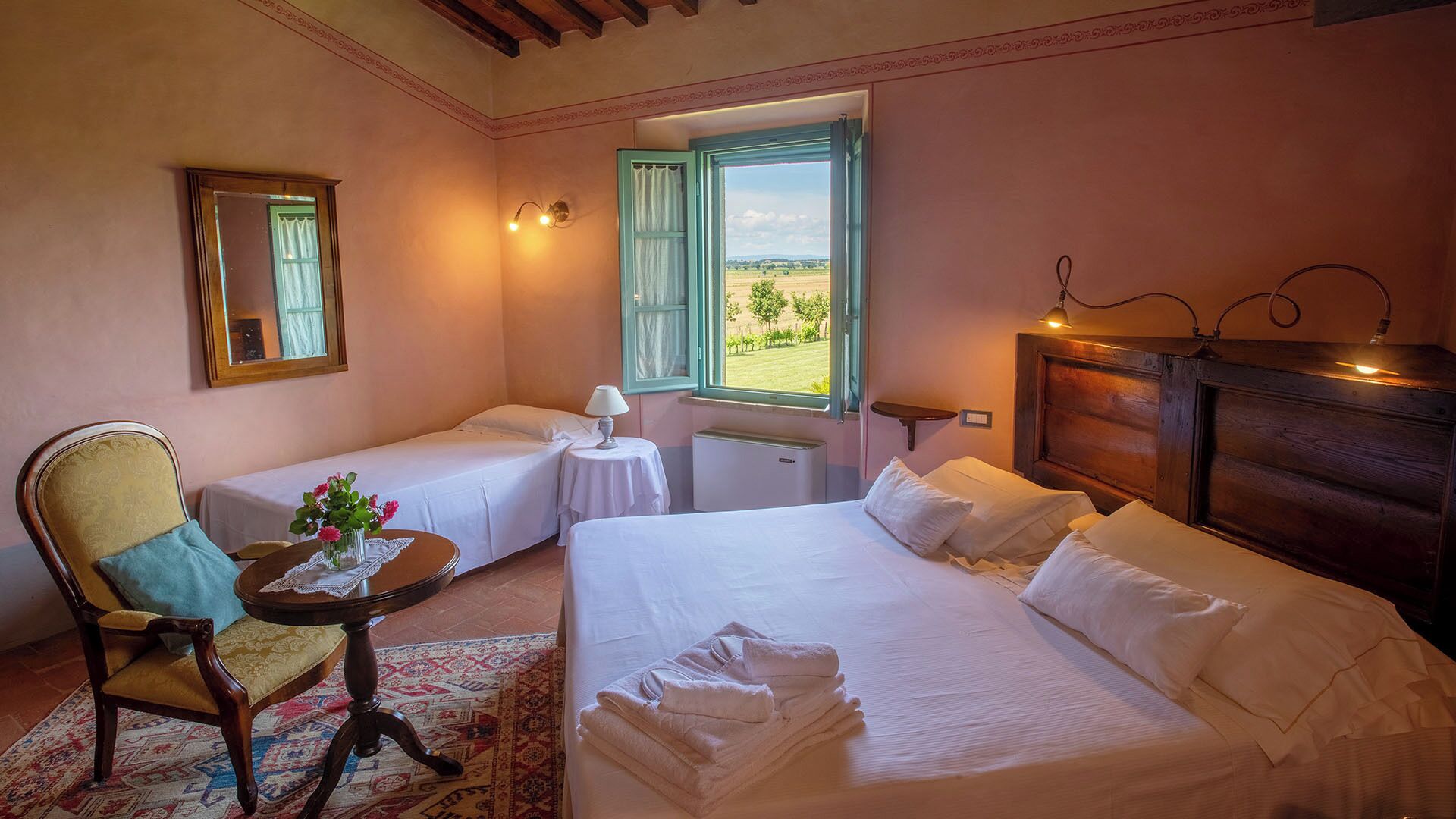 A hotel room with two single beds, a wooden headboard, a small table with a lamp, a chair, and a view of the countryside through an open window.