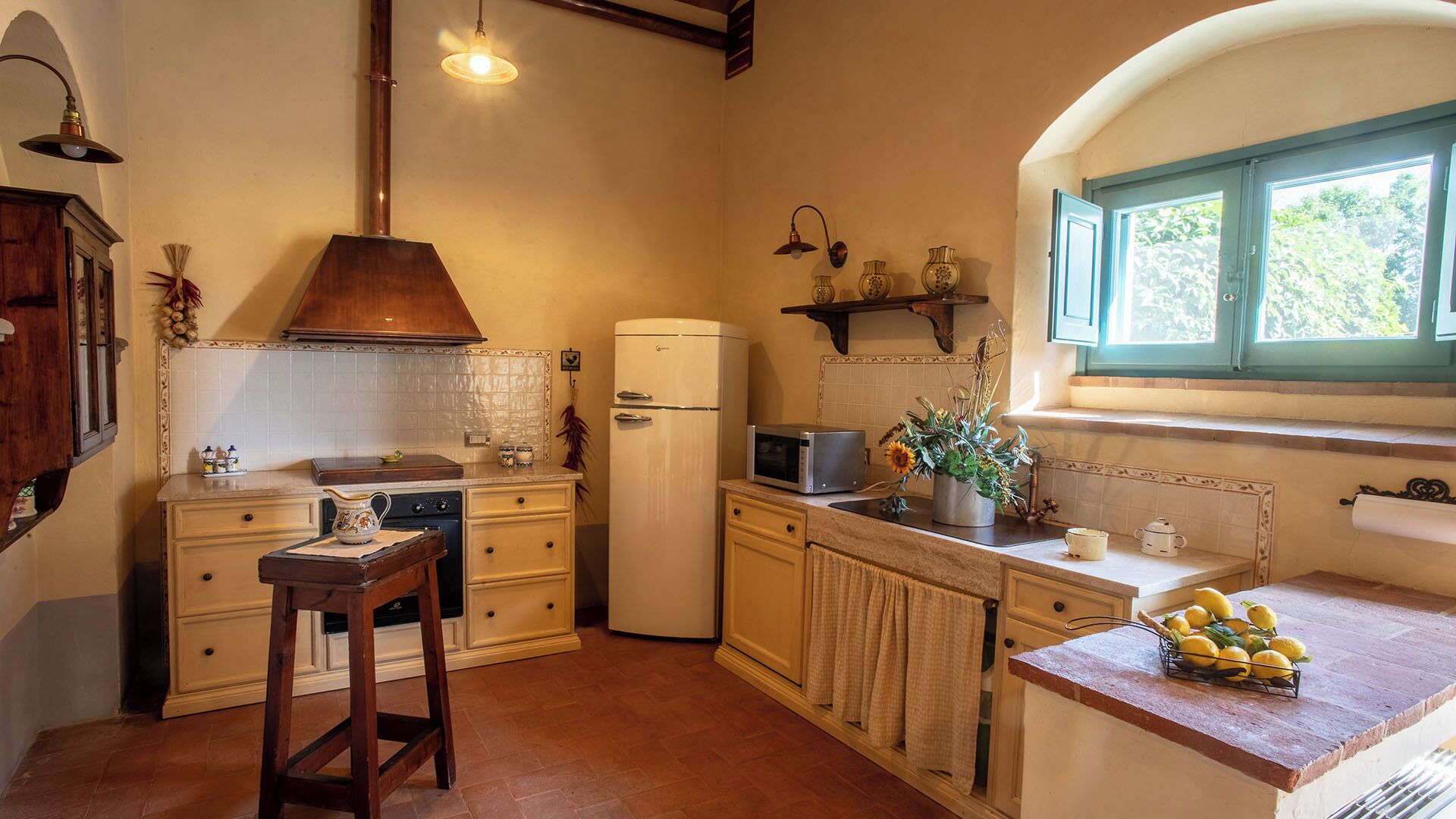 A kitchen with a wooden stool, a white refrigerator, a microwave, and a window with a view of greenery.