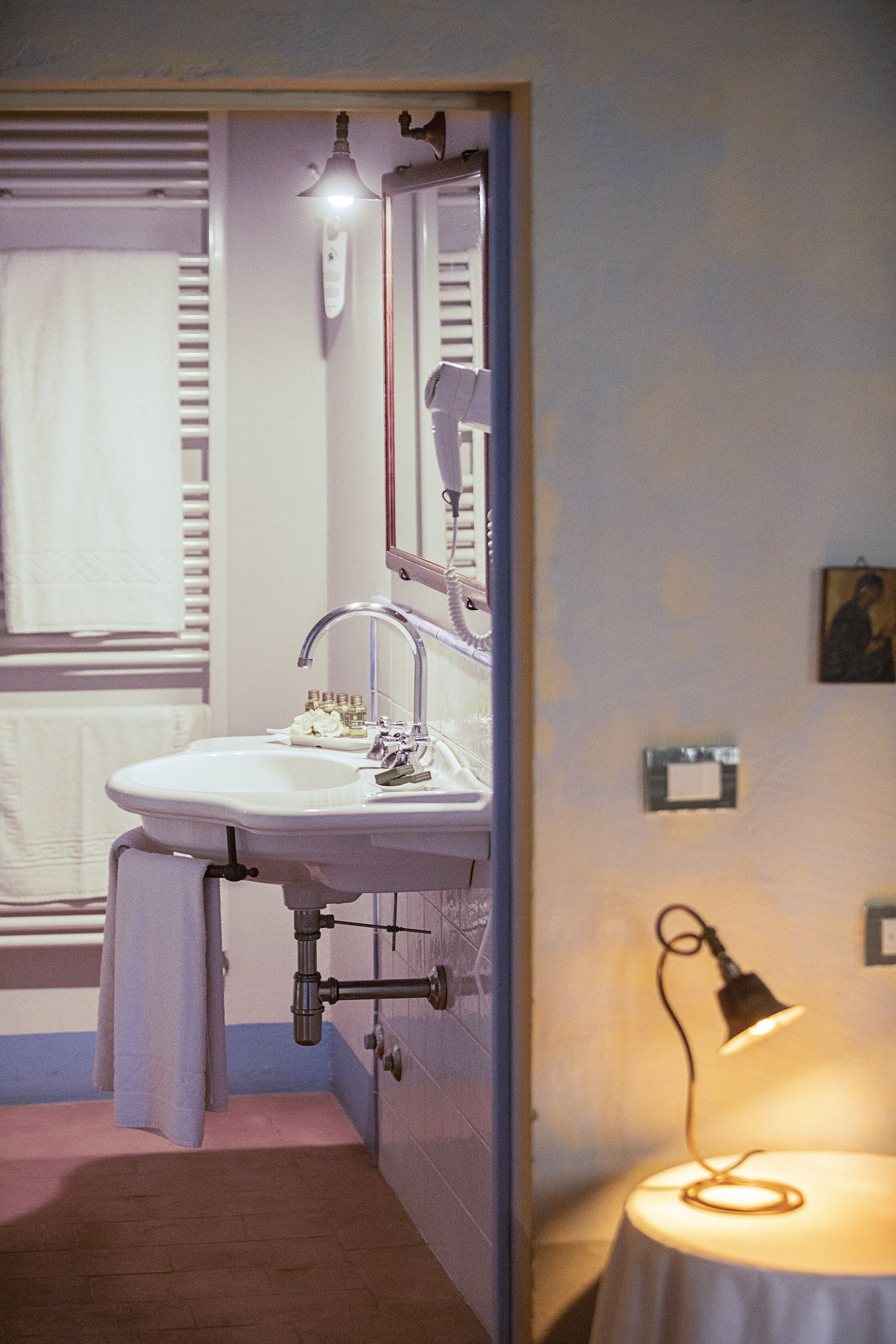 Un baño con lavamanos blanco, espejo y barra para toallas. Hay una mesita con una lámpara y un cuadro en la pared.