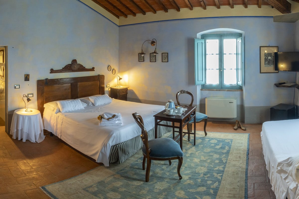 Un dormitorio con techo de madera, una cama con ropa blanca, una mesita con una silla, una ventana con cortinas y un televisor en la pared.
