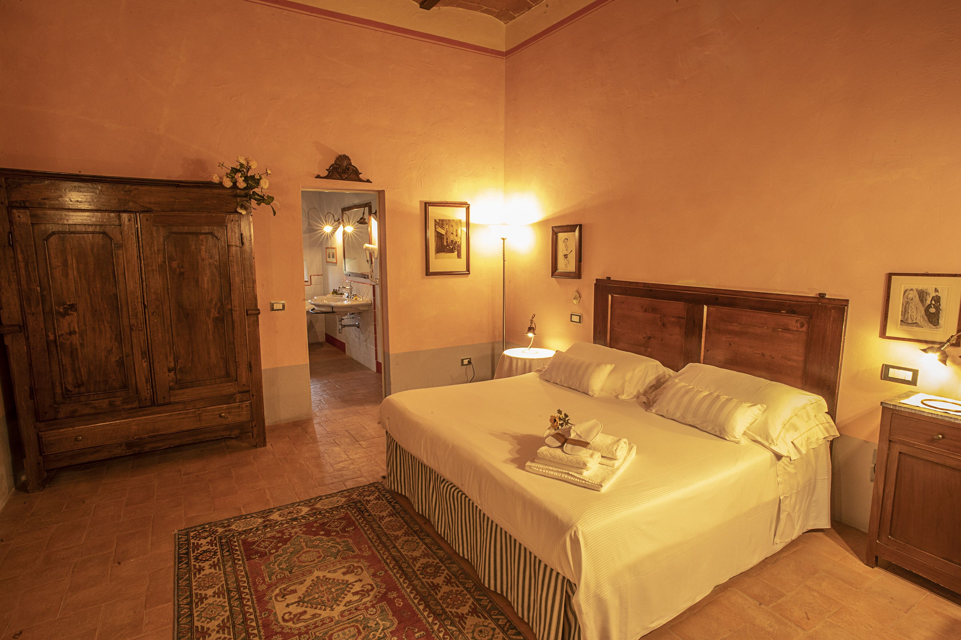 A bedroom with a large wooden wardrobe, a bed with white linens, a bedside table with a lamp, and a rug on the floor.