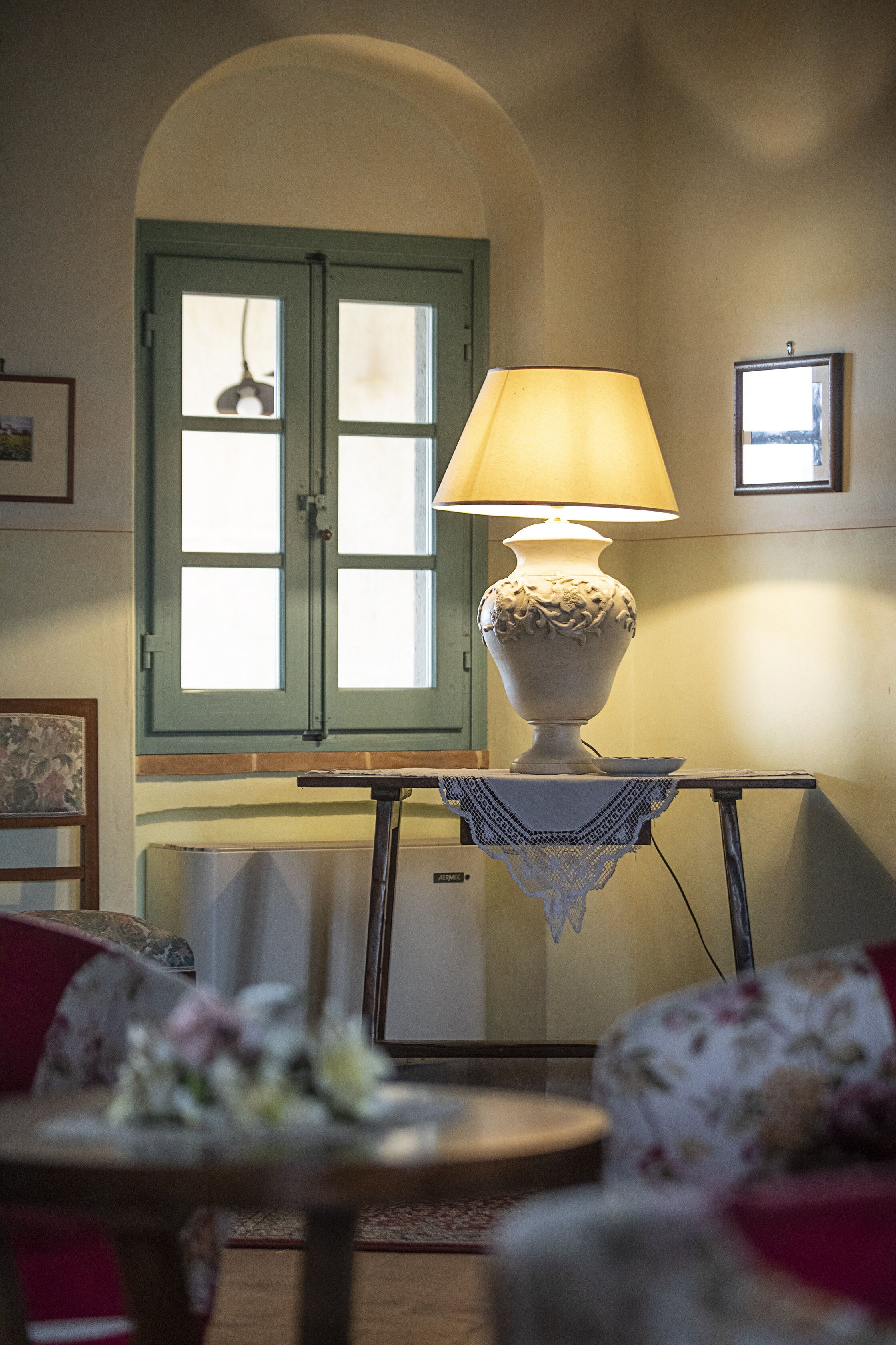 Ein Zimmer mit einer Tischlampe, einem Fenster mit grünem Rahmen und einem kleinen Tisch mit weißer Spitzenleinentuch.