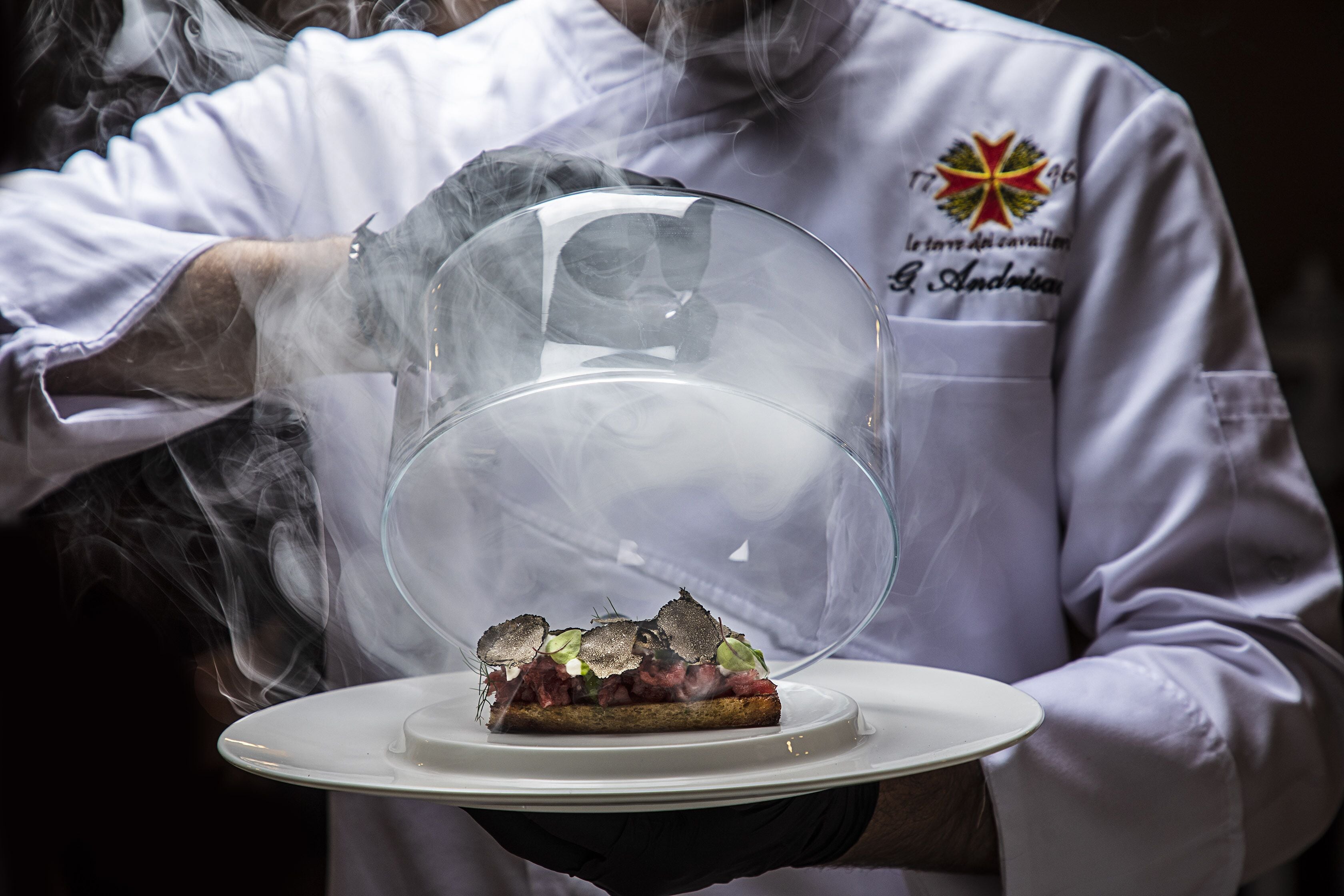 A person in a white chef's uniform holding a plate with a dish covered by a transparent dome.