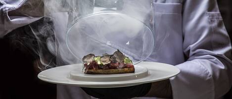 A person in a white chef's uniform holding a plate with a dish covered by a transparent dome.