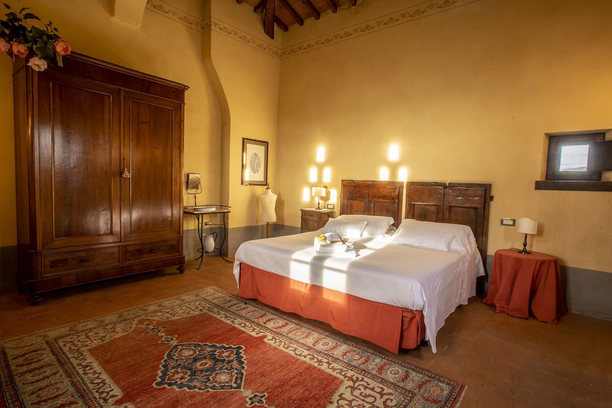 Habitación de hotel con un amplio armario de madera, cama con ropa de cama blanca, alfombra estampada y una pequeña mesa con lámpara.