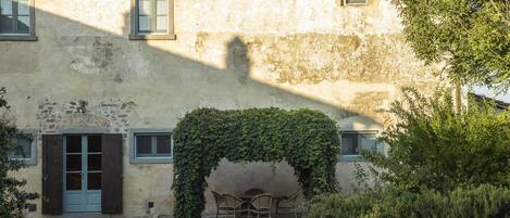 Un edificio con fachada rústica, una puerta con contraventanas y un pequeño espacio al aire libre bajo un arco cubierto de hiedra.