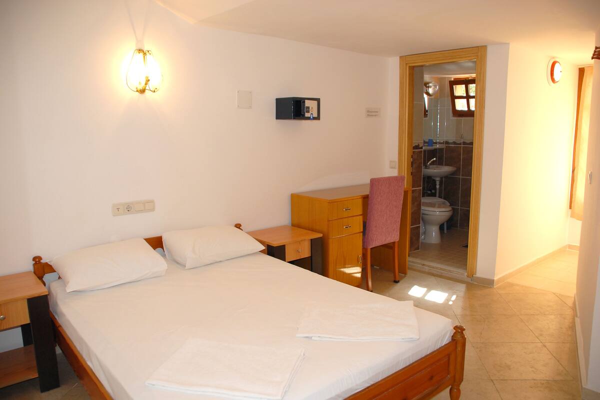 A bedroom with a bed, bedside table, desk, chair, and a bathroom visible through an open door.