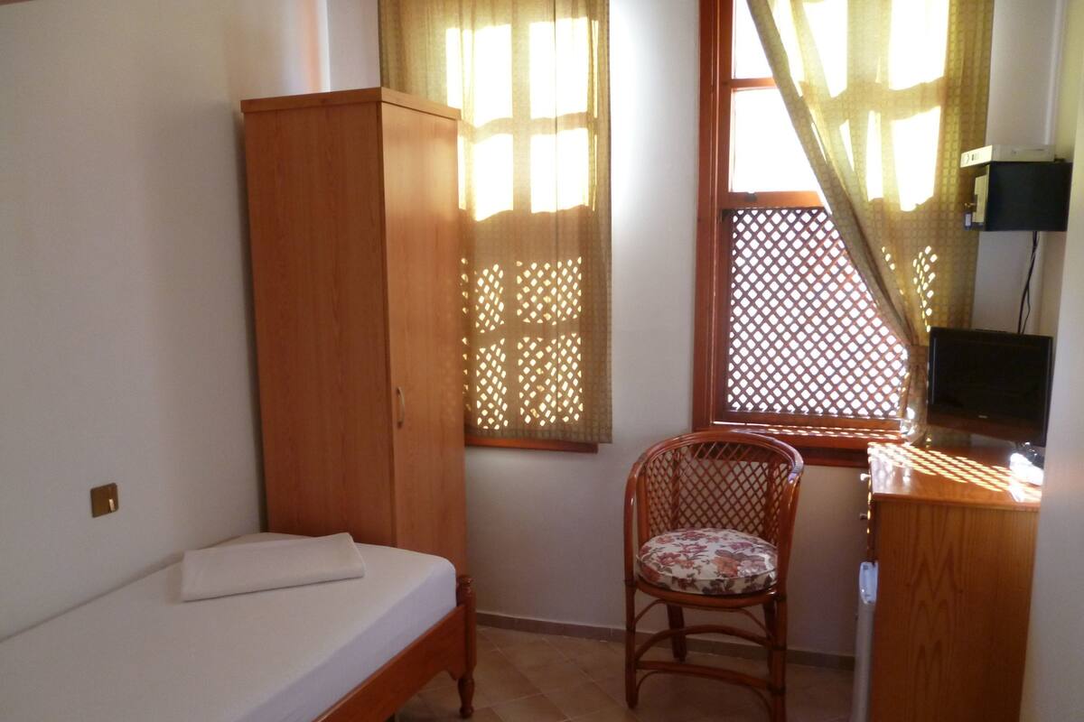 A small, well-lit room with a single bed, a wooden wardrobe, a wicker chair, a desk with a television, and a window with a lattice screen.