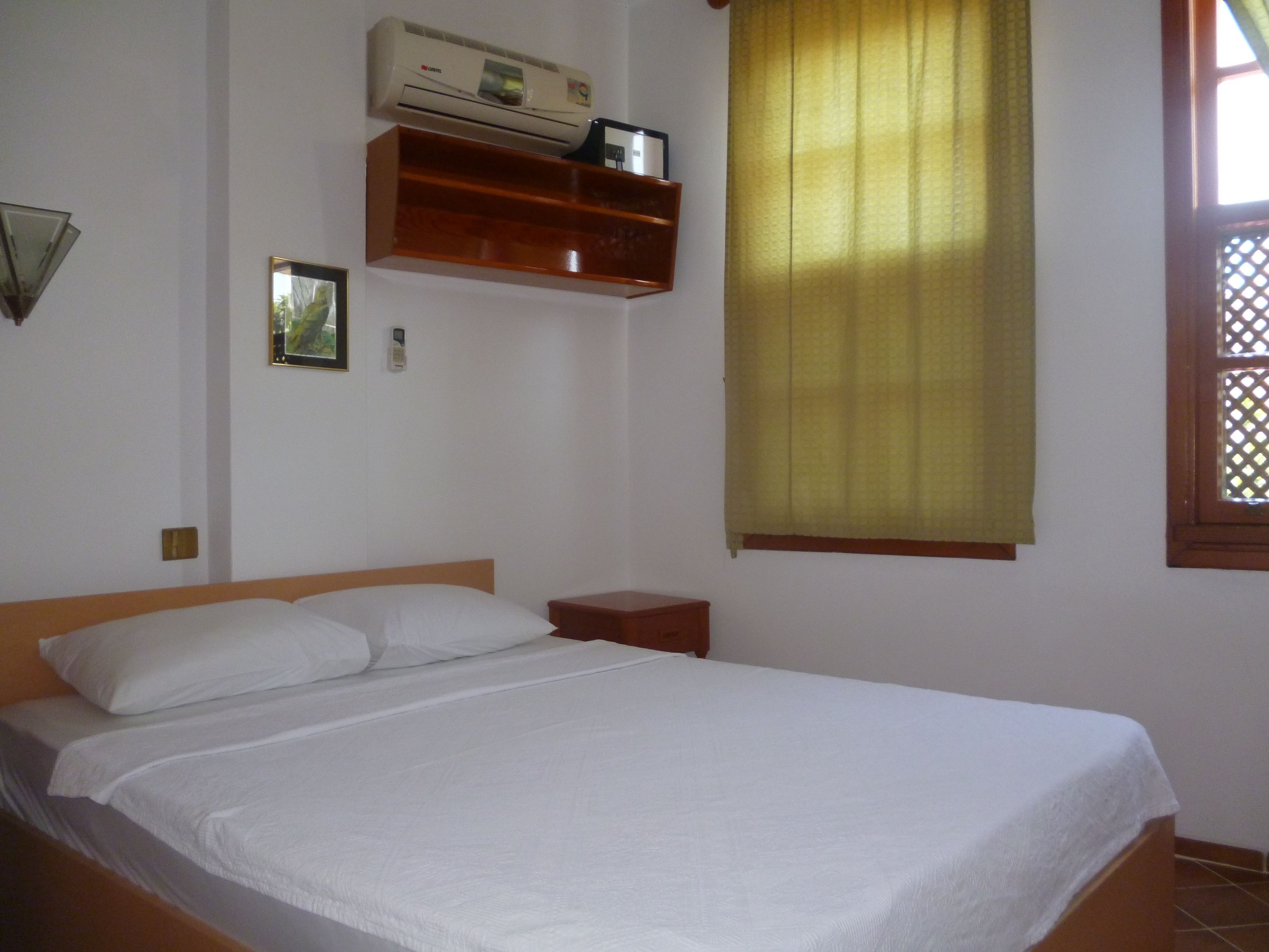 A single bed with white bedding, a wooden headboard, a small bedside table, a wall-mounted shelf with a clock, and a window with yellow curtains.