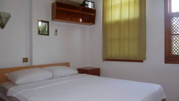 A single bed with white bedding, a wooden headboard, a small bedside table, a wall-mounted shelf with a clock, and a window with yellow curtains.