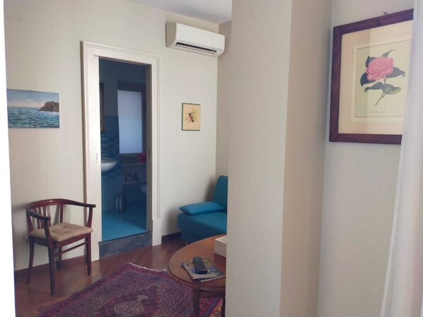 A room with a wooden chair, a blue sofa, a small table with a remote control, a framed picture of a rose, and an air conditioning unit on the wall.