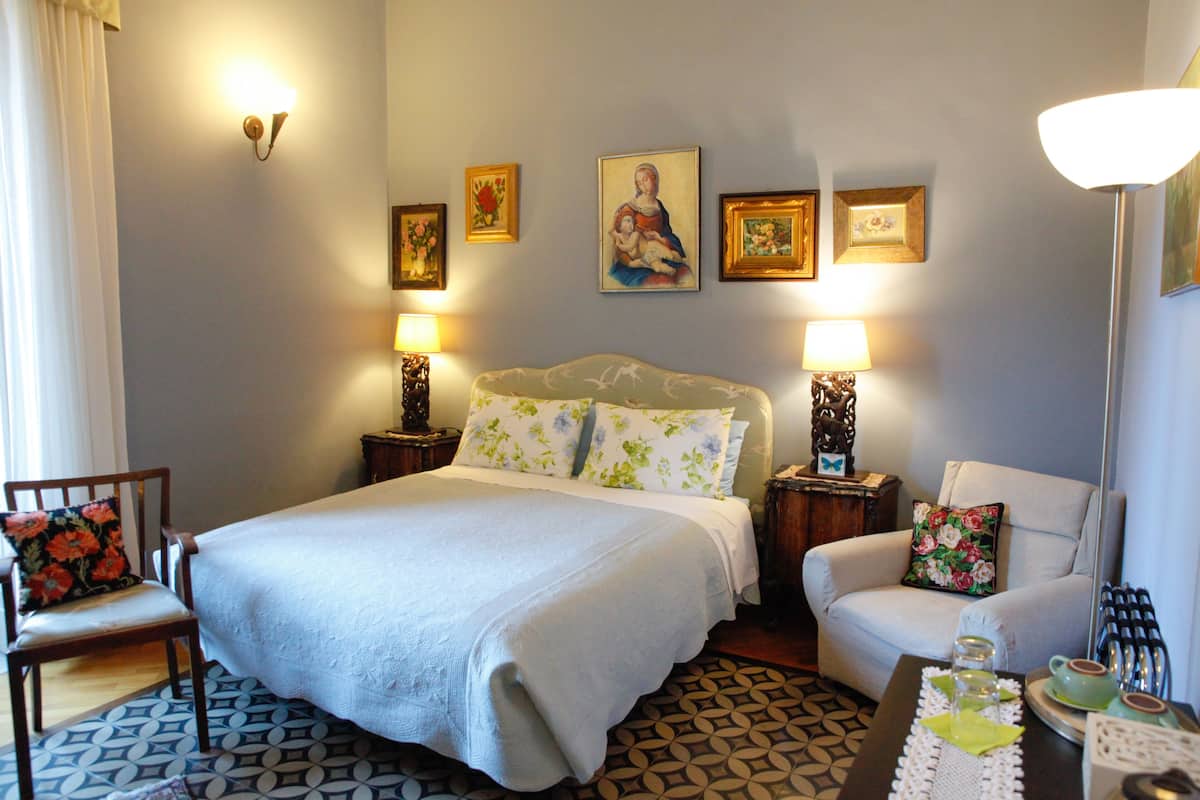 Habitación de hotel con una cama grande, dos mesitas de noche, una silla y un sofá. Cuenta con lámparas de pared, cuadros enmarcados y un suelo estampado.