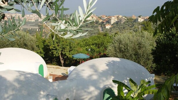 Vista de una zona costera con estructuras blancas en forma de cúpula, probablemente alojamientos, rodeada de vegetación y con vistas a un paisaje urbano.