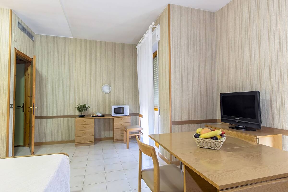 A compact room with a bed, a desk with a computer, a television, and a small dining table with fruit.