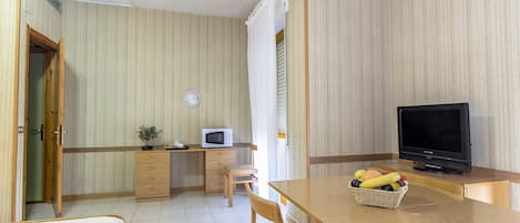 A compact room with a bed, a desk with a computer, a television, and a small dining table with fruit.
