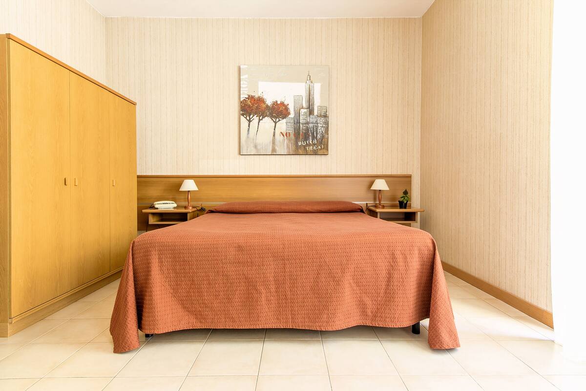 A hotel room with a large bed, wooden wardrobe, two bedside tables with lamps, and a cityscape artwork on the wall.