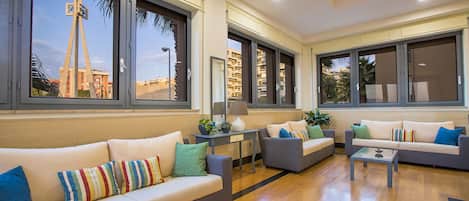 Un salón moderno con un sofá beige, una mesa de centro de cristal y un sillón gris. Grandes ventanales ofrecen vistas a edificios cercanos.