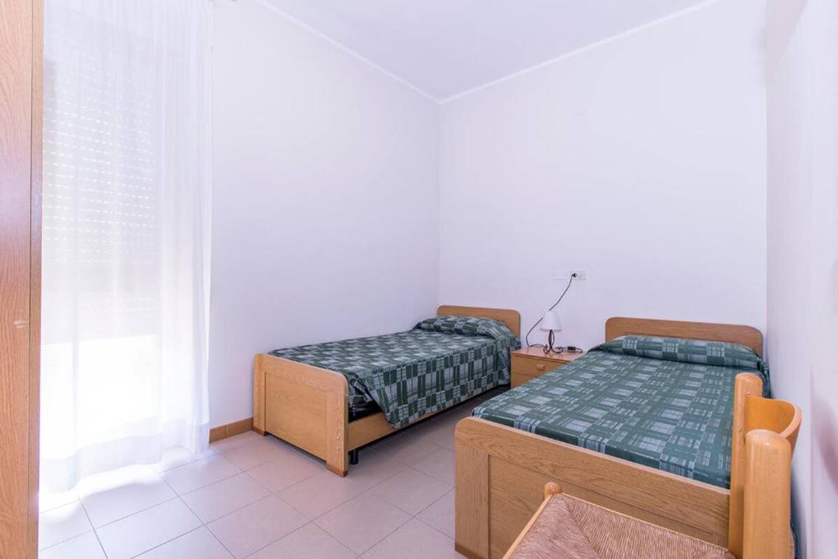 A small, clean room with two wooden beds, a small bedside table, and a window with sheer curtains.