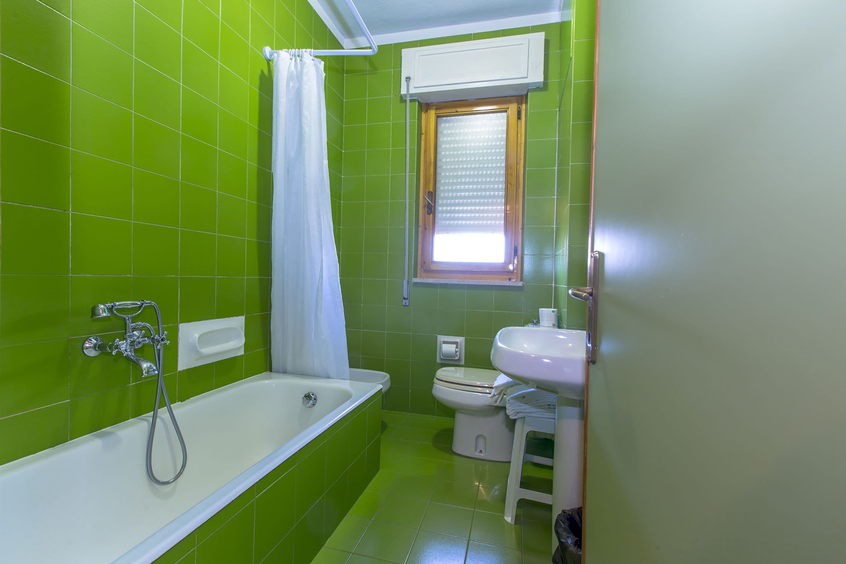A bathroom with green tiles, a white bathtub, a toilet, and a window with blinds.
