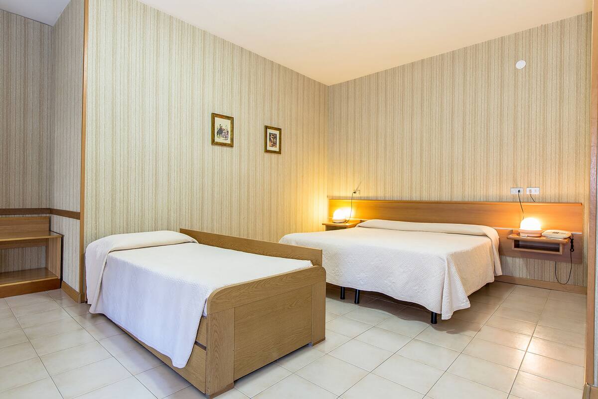 A hotel room with two beds, a wooden bench, and a nightstand with a lamp.