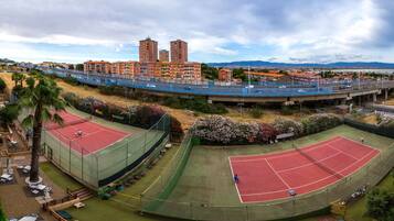Una cancha de tenis con una valla perimetral, una palmera y vistas de edificios al fondo.