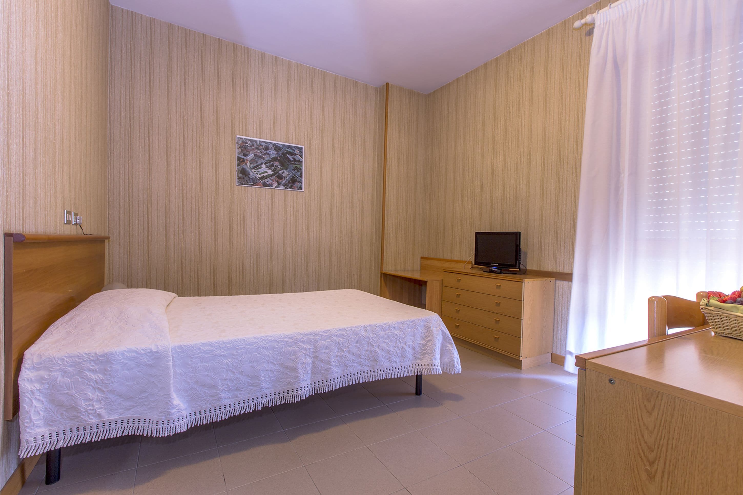 Ein ordentlich eingerichtetes Schlafzimmer mit einem Bett, einer Holzkommode, einem Fernseher und einem Schreibtisch mit Korb.