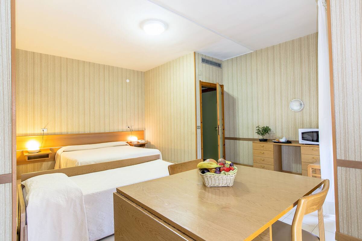 A hotel room with two beds, a dining table with a fruit basket, a microwave, and a small desk with a plant.