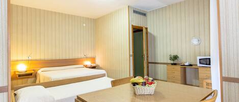 A hotel room with two beds, a dining table with a fruit basket, a microwave, and a small desk with a plant.