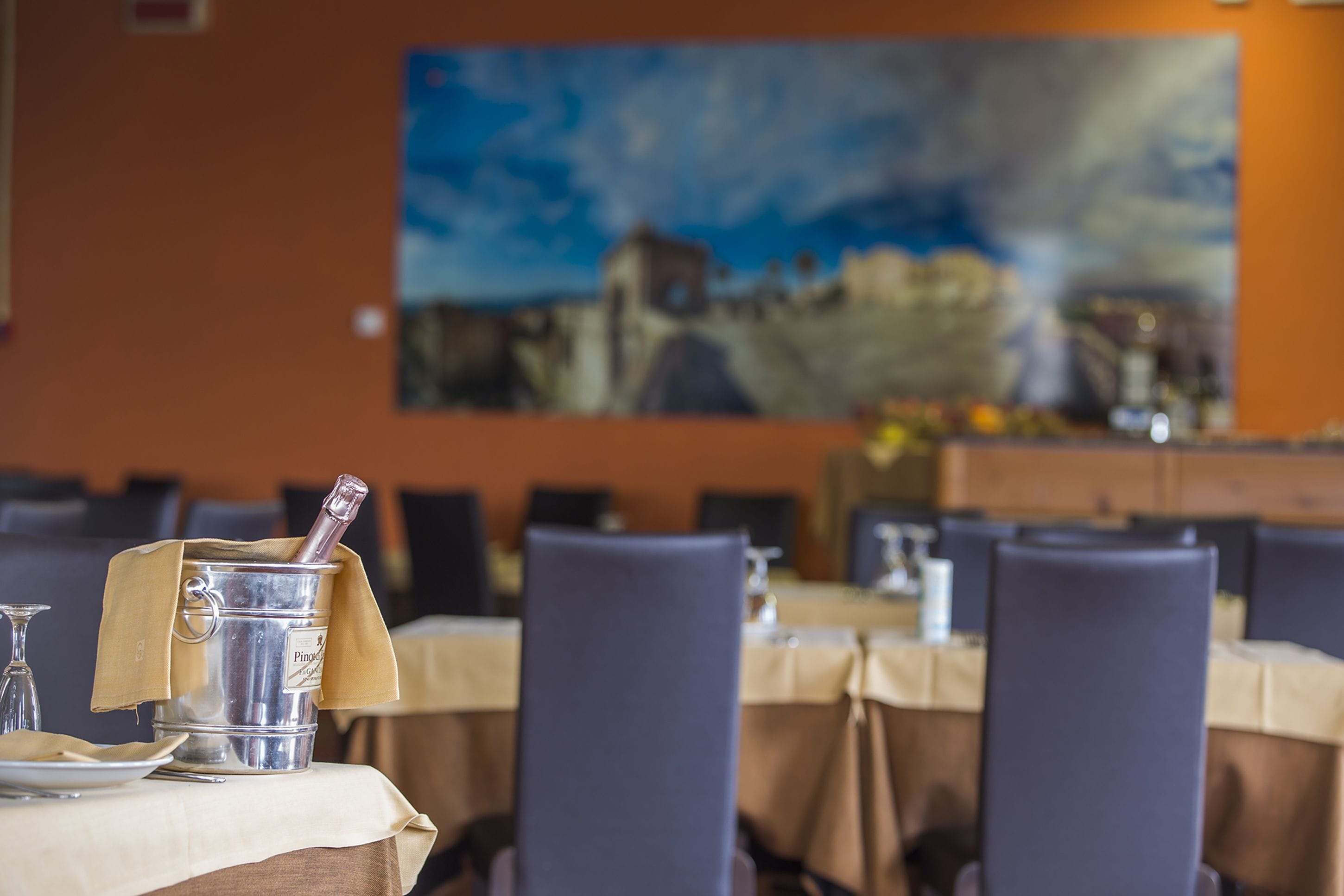 A dining area with tables set for service, a bottle of champagne, and a painting on the wall.