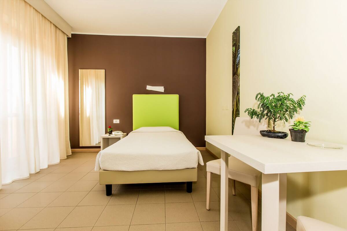 A hotel room with a bed, a desk, and a chair. There is a nightstand with a lamp and a potted plant.