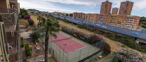 A tennis court surrounded by greenery and a residential area with high-rise buildings in the background.