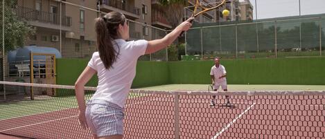 A tennis court with a net, a player in a white shirt and striped shorts, and a palm tree in the background.