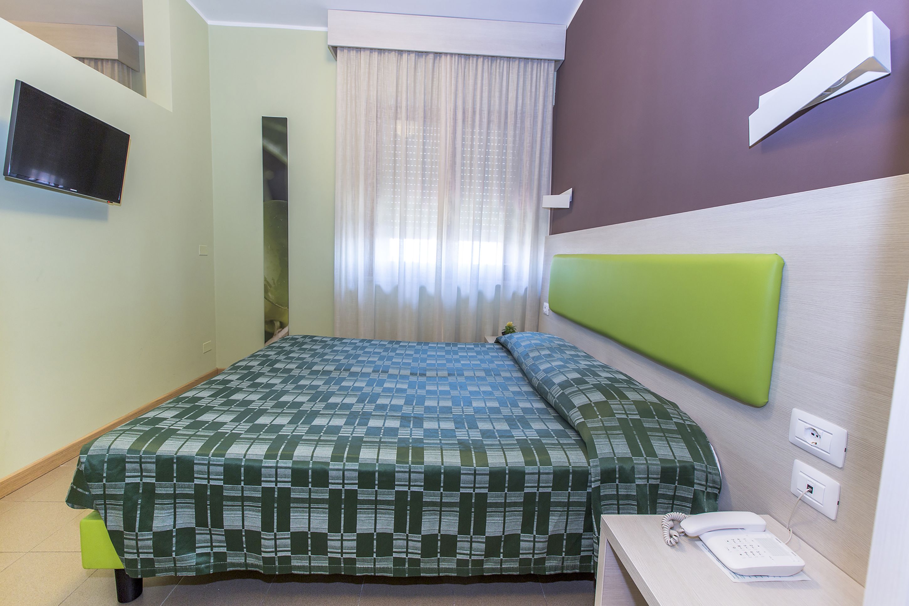 Hotellihuone, jossa on sänky, yöpöytä, jossa on puhelin, seinälle asennettu televisio ja ikkuna, jossa on verhot.