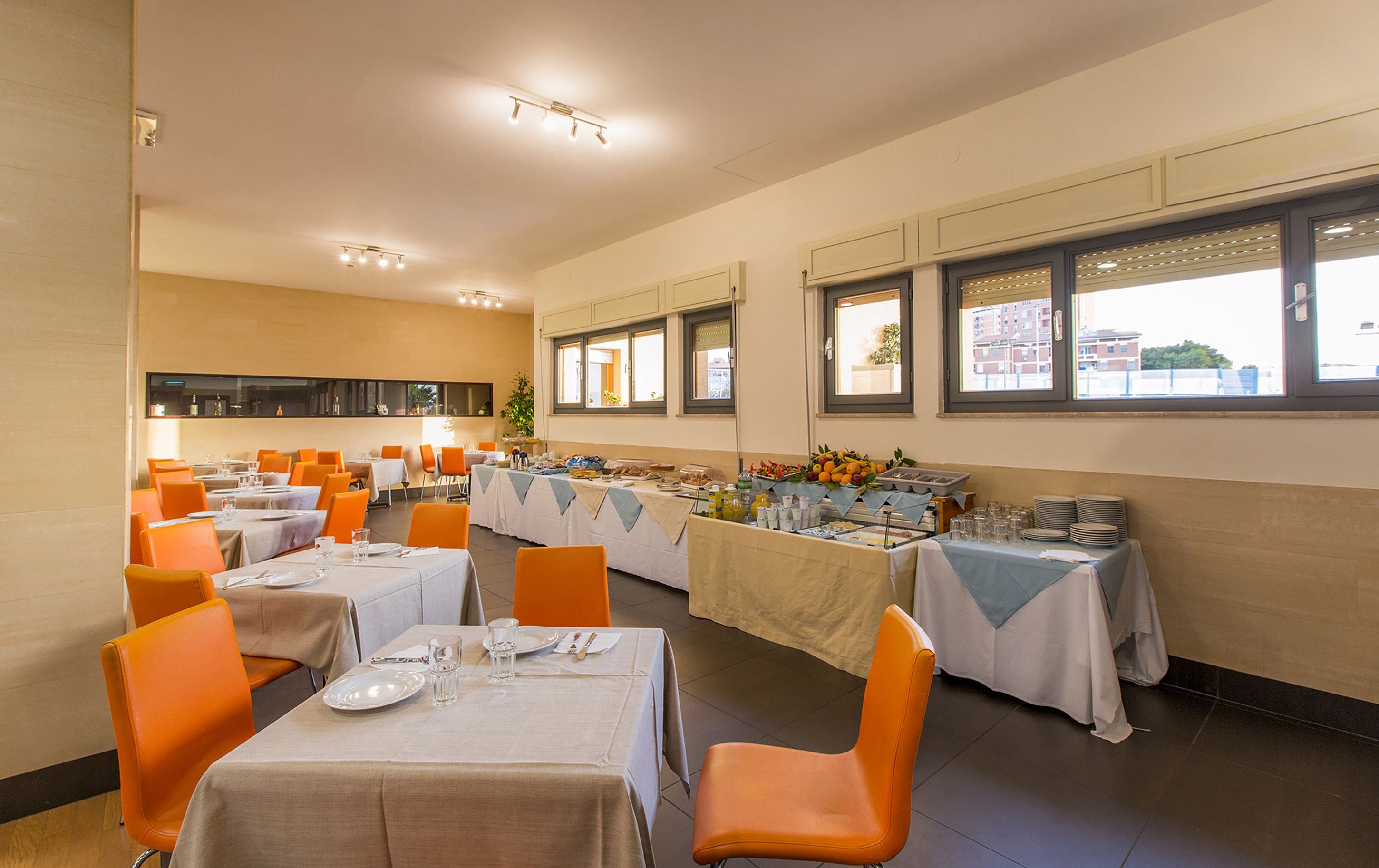 A dining area with multiple tables set for meals, orange chairs, and a buffet with various food items.