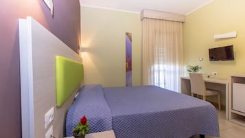A hotel room with a bed, a desk, a chair, a TV, and a plant.