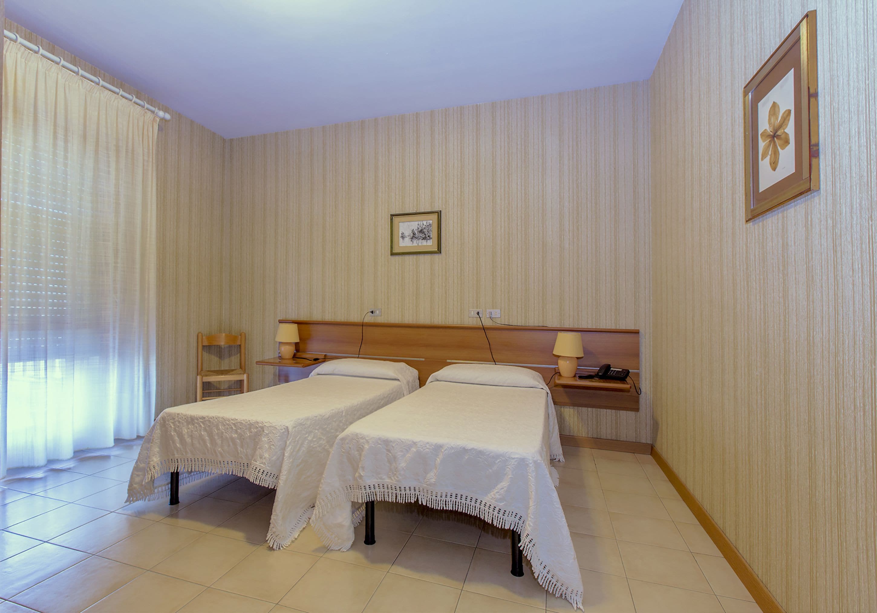 Une chambre d’hôtel avec deux lits, une table de chevet avec une lampe, un tableau encadré au mur et une fenêtre avec des rideaux.