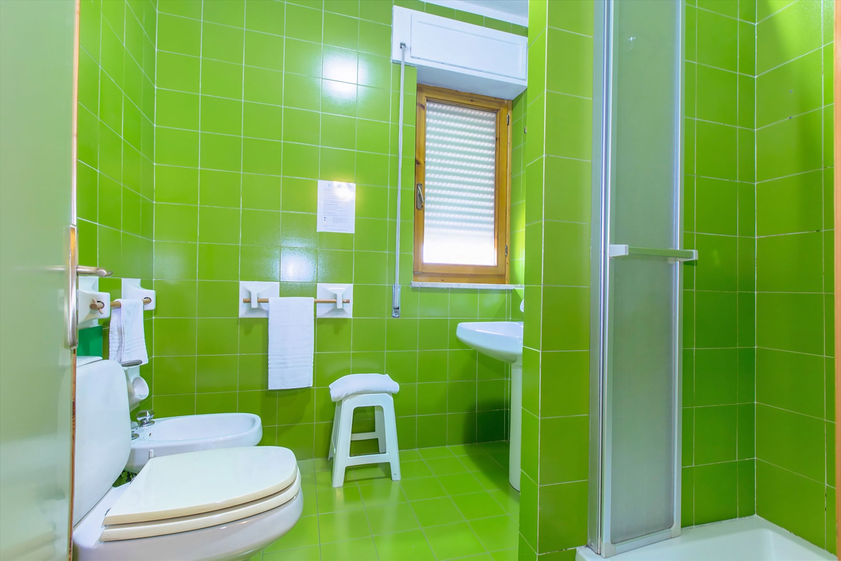 Un baño con azulejos verdes, un inodoro, un lavabo, una ducha y un taburete pequeño.