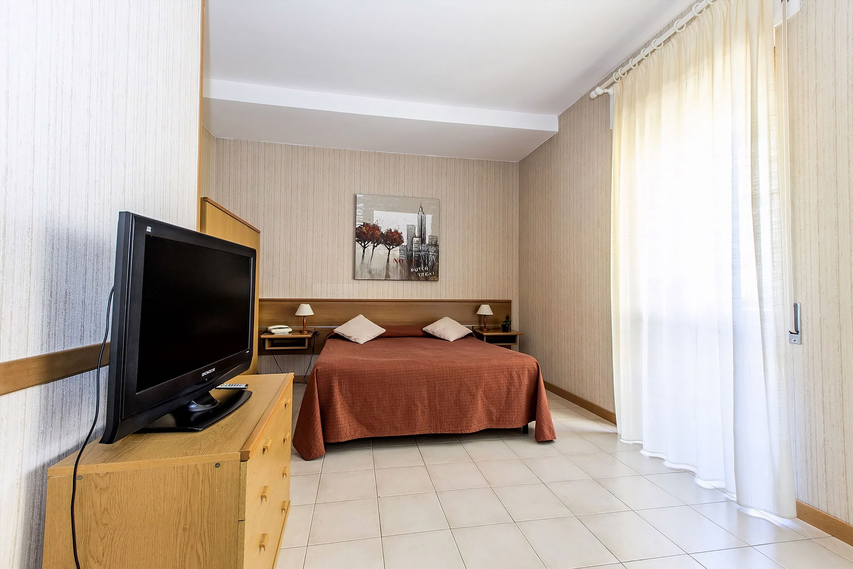 A hotel room with a bed, a television on a wooden stand, a nightstand with a lamp, and a painting on the wall.