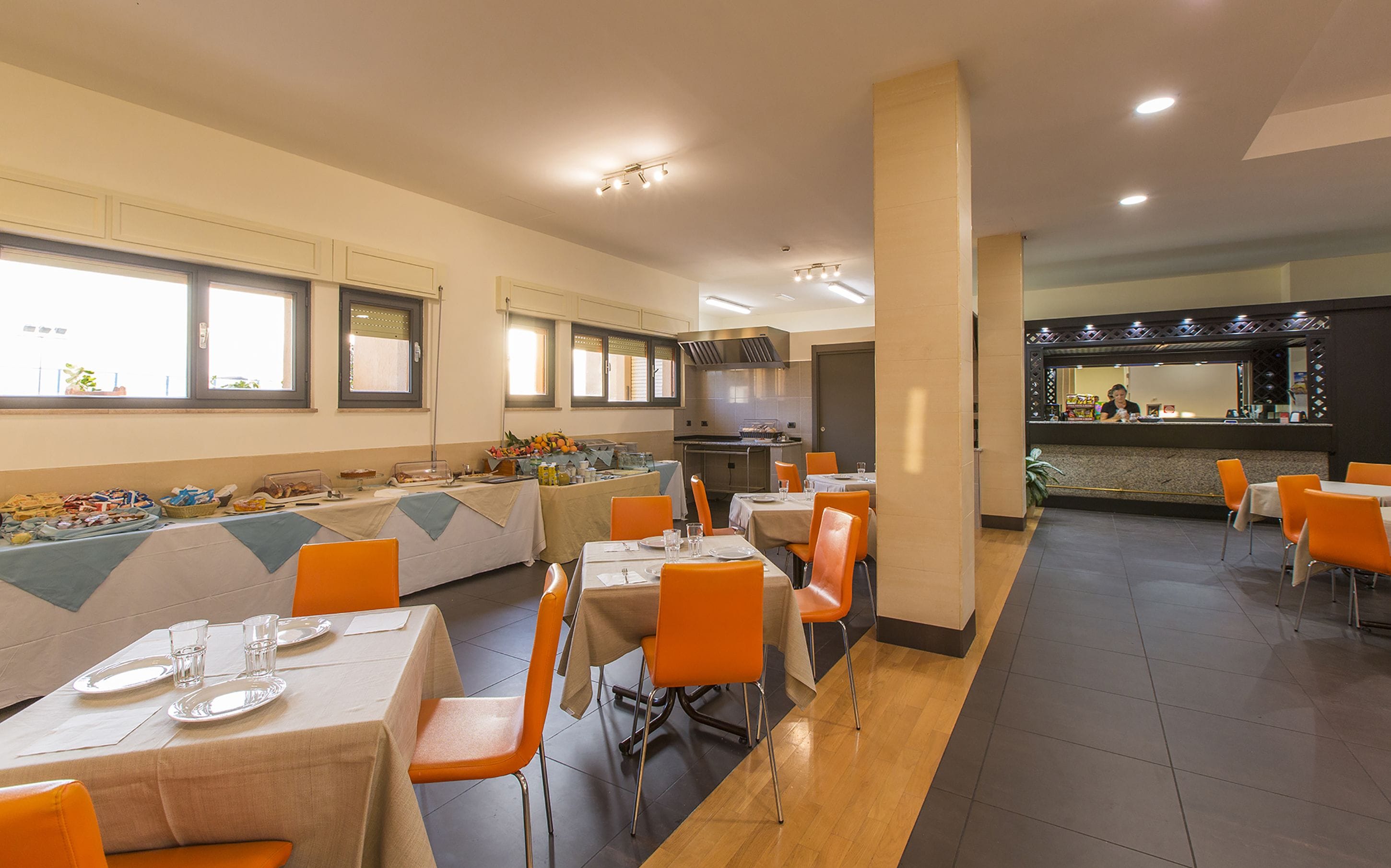 A buffet-style dining area with multiple tables and orange chairs, a kitchen in the background, and a bar area with a counter and stools.