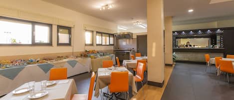 A buffet-style dining area with multiple tables and orange chairs, a kitchen in the background, and a bar area with a counter and stools.