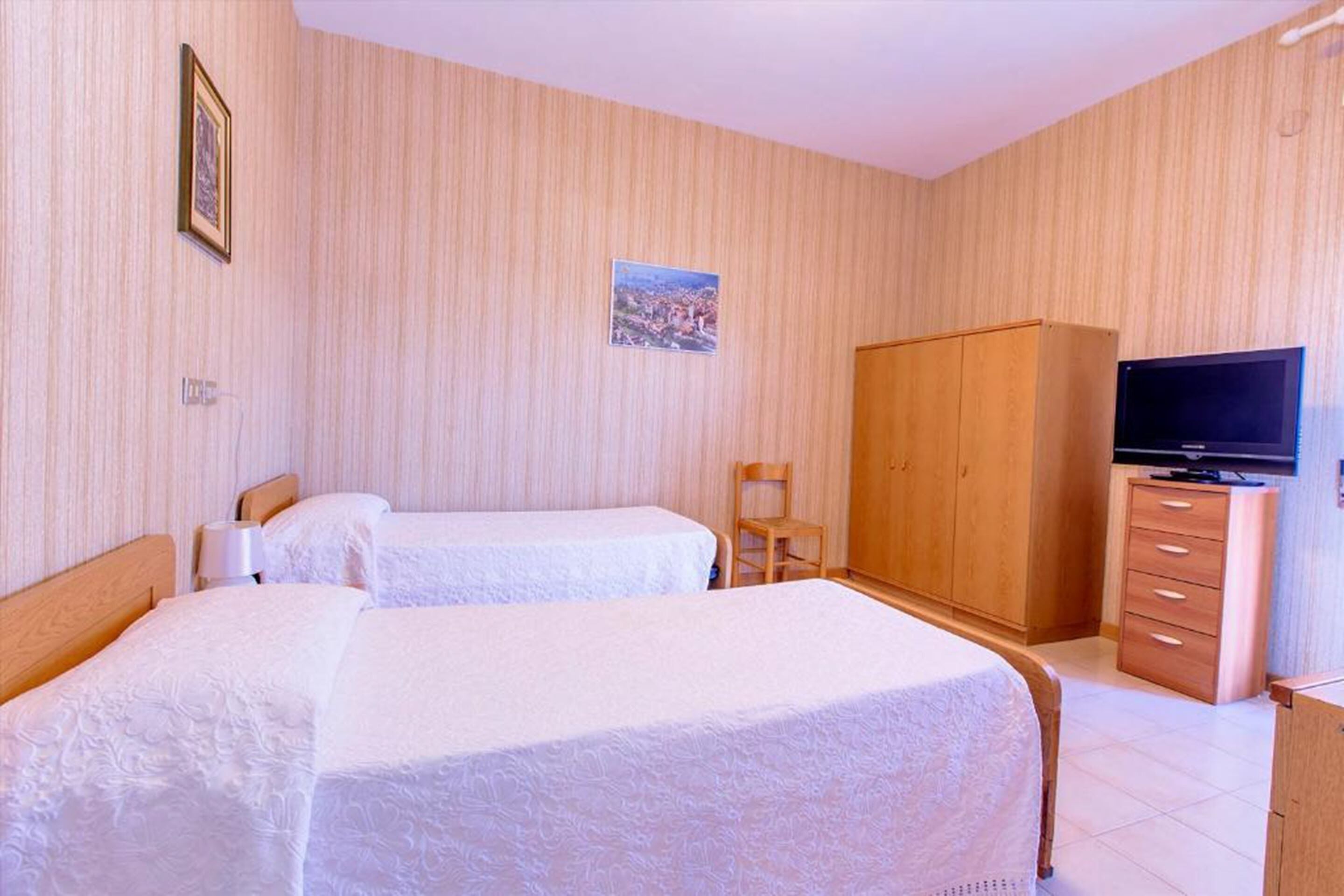 A hotel room with two beds, a wooden wardrobe, a television, and a small chair.