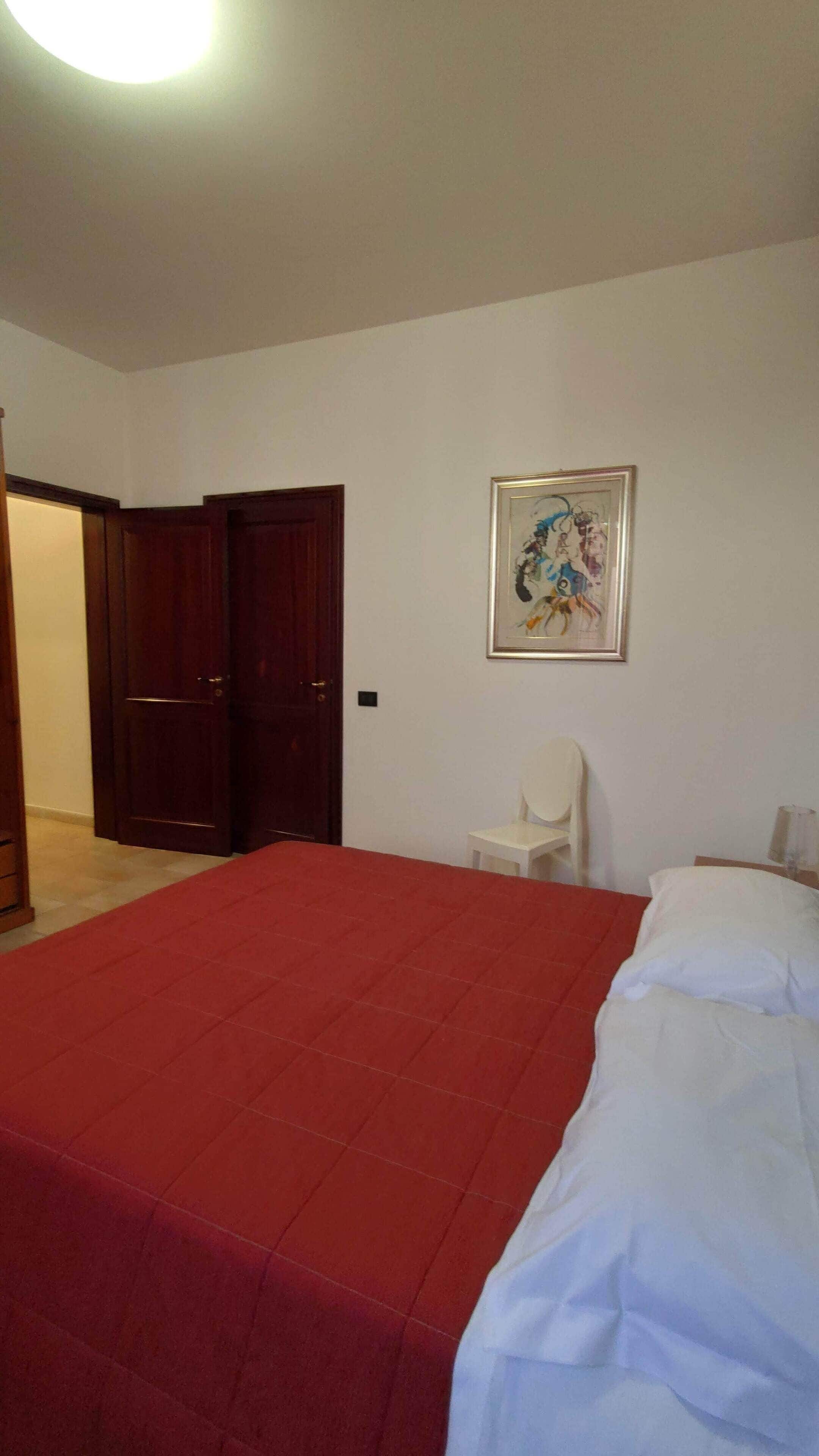 A bedroom with a red bedspread, a white bed, a framed painting on the wall, and a white chair.