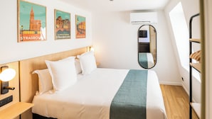 Una cama bien hecha con ropa blanca y un plaid verde, una mesita de noche con lámpara, y dos carteles de Estrasburgo en la pared.