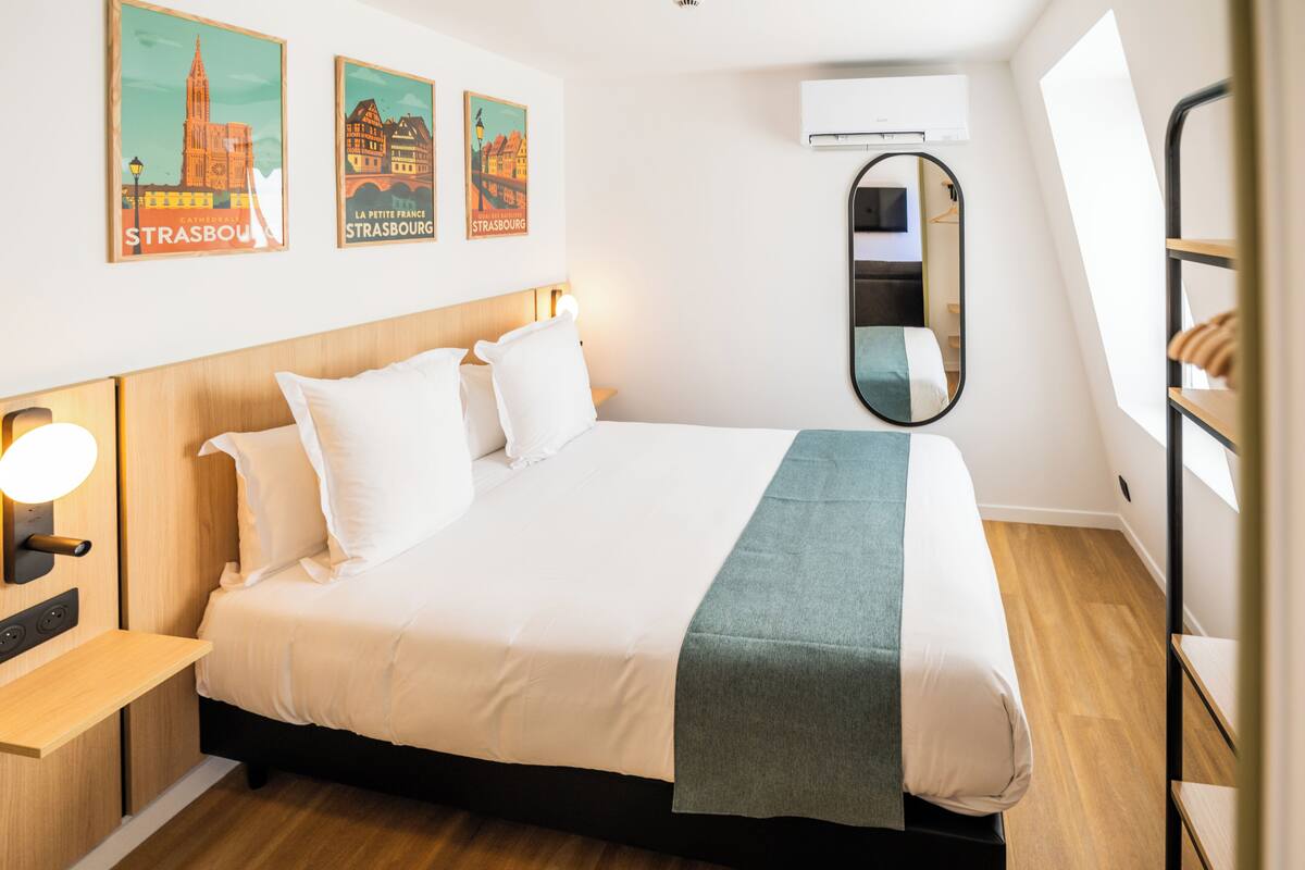 A neatly made bed with white bedding and a green throw, a bedside table with a lamp, and two posters of Strasbourg on the wall.