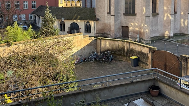 Un patio con un edificio que tiene ventanas arqueadas y un techo de tejas, rodeado de árboles y bicicletas estacionadas.