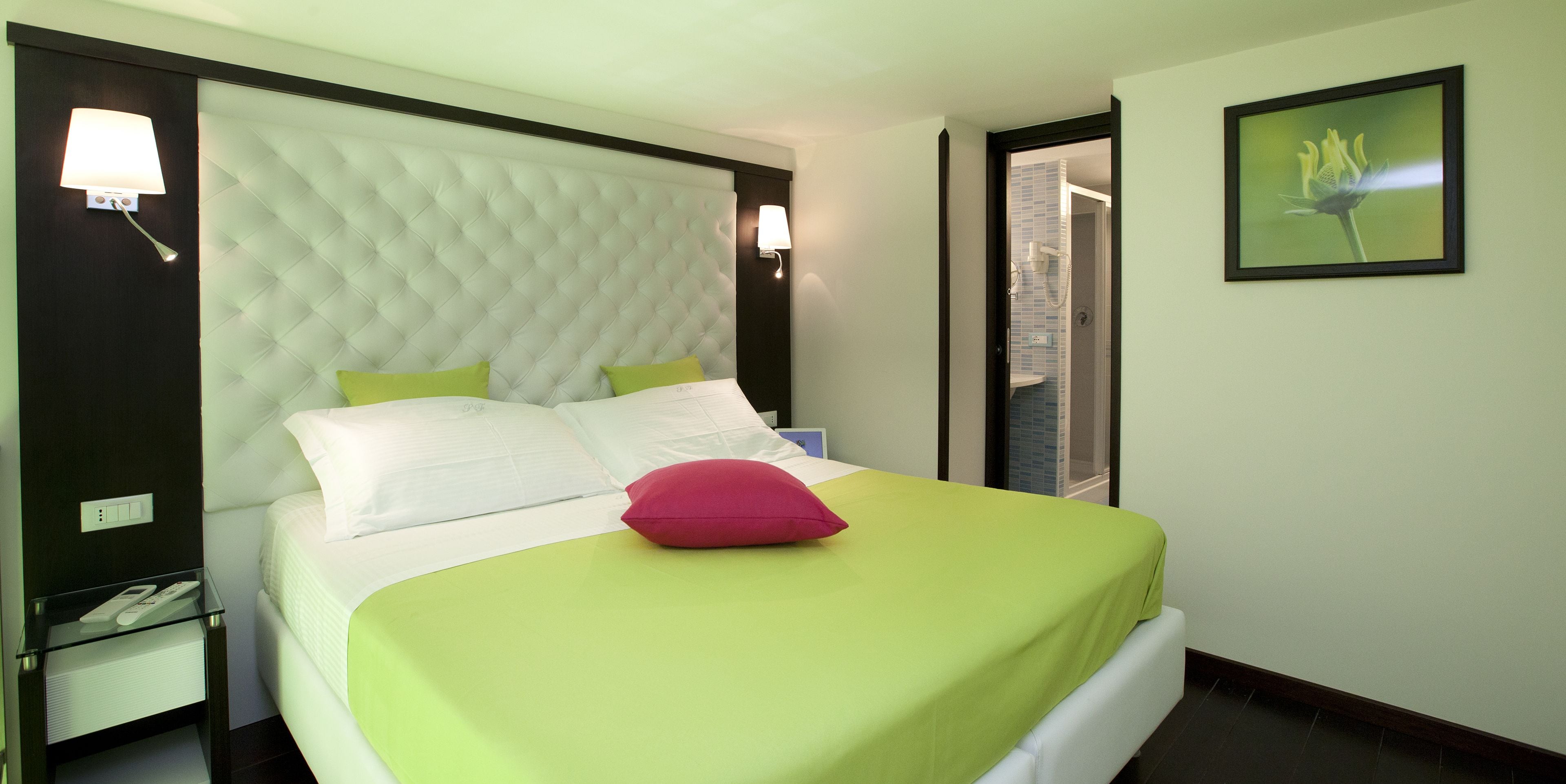 A hotel room with a tufted headboard, a bed with white and green bedding, a red pillow, a bedside table with a lamp, and a framed picture on the wall.