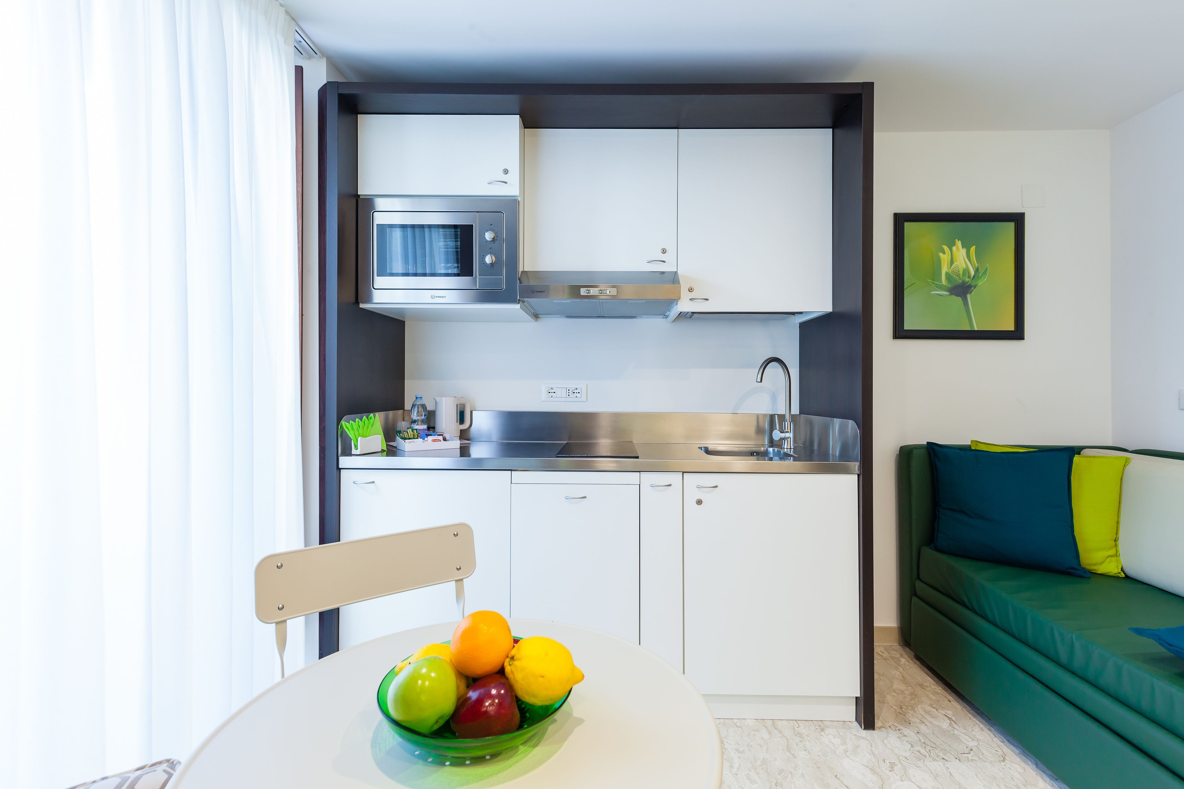 Une cuisine compacte avec des meubles blancs, un four à micro-ondes et un évier. On y trouve une table à manger avec un saladier de fruits et une chaise. Un canapé vert agrémenté de coussins colorés se trouve dans l’espace salon.