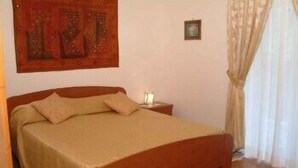 Un dormitorio con cama de madera, ropa de cama color beige, una mesita de noche y un pequeño tocador, además de un cuadro en la pared.
