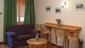 Una sala pequeña con un sofá, una mesa de comedor con sillas, una mesita redonda con un objeto decorativo y una ventana con cortinas verdes.